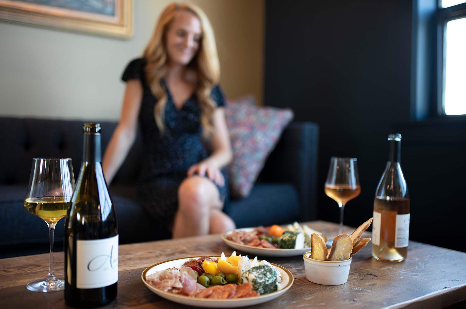 Young woman relaxing, blurred in the background, Anaba wine and food platters placed in the foreground in front of her. 