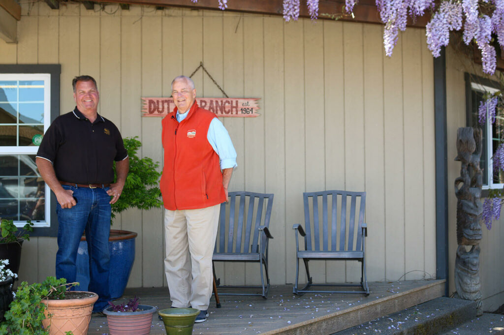 Two smiling men positioned in front of Dutton Ranch on a porch with plant pots on the left, and two chairs on the right. 