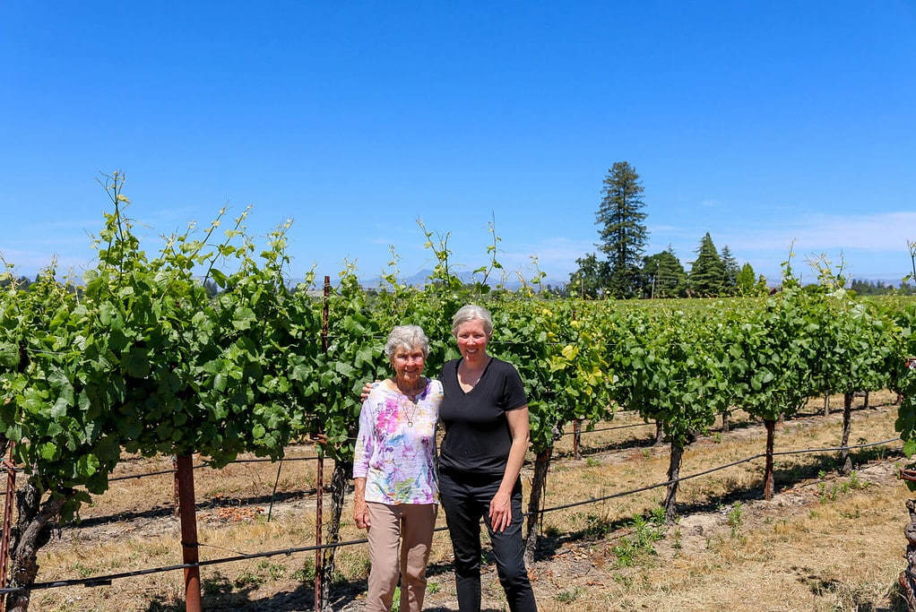 Two women smiling in front of a row of blooming vines at O'Connor Vineyard, a cluster of trees to the right in the background, under a clear blue sky. 