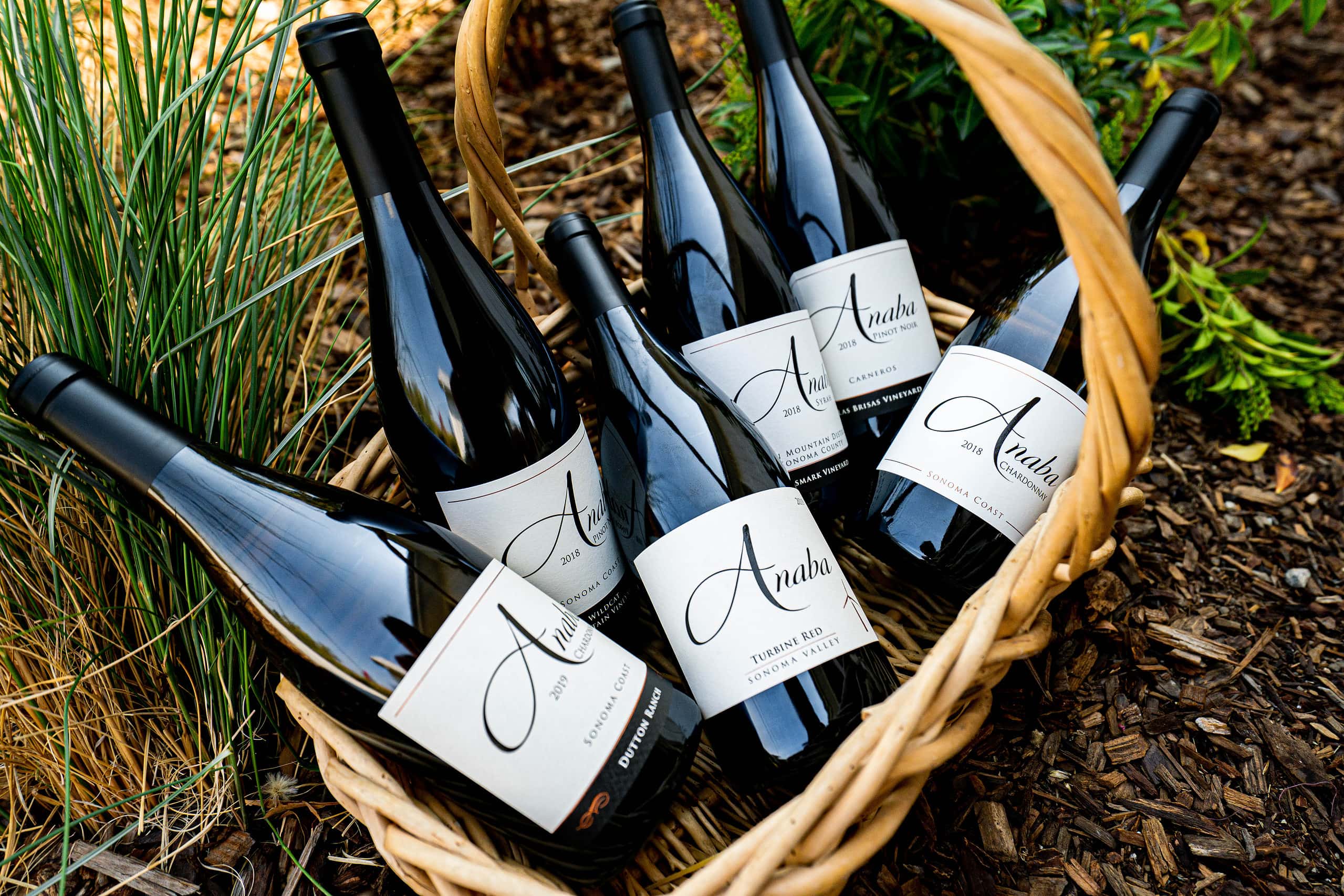 Six bottles of wine are placed in a wooden basket next to grass.
