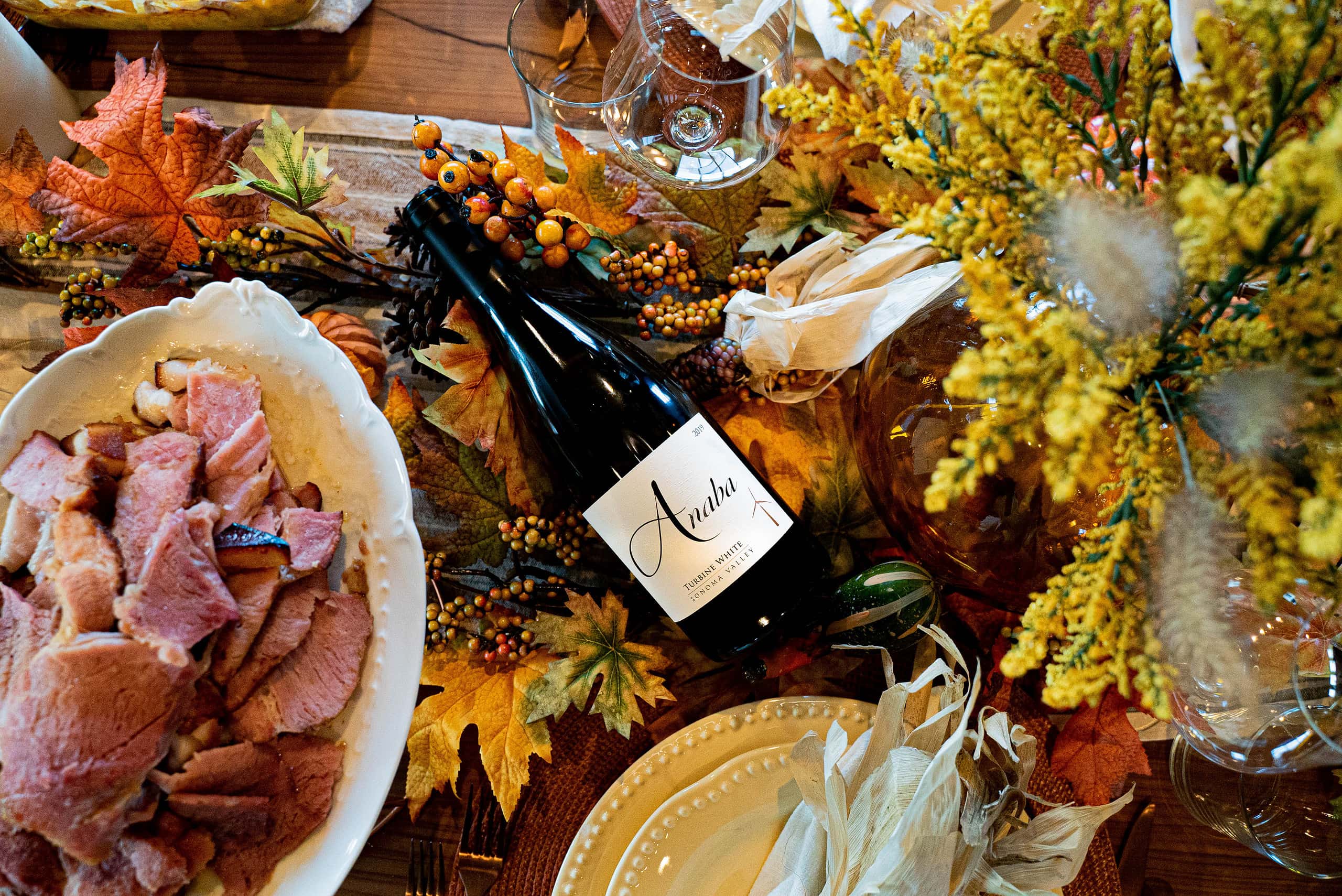 A thanksgiving dinner table is decorated with fall leaves and flowers, with a platter of ham, a bottle of Anaba's Turbine White is in the middle.