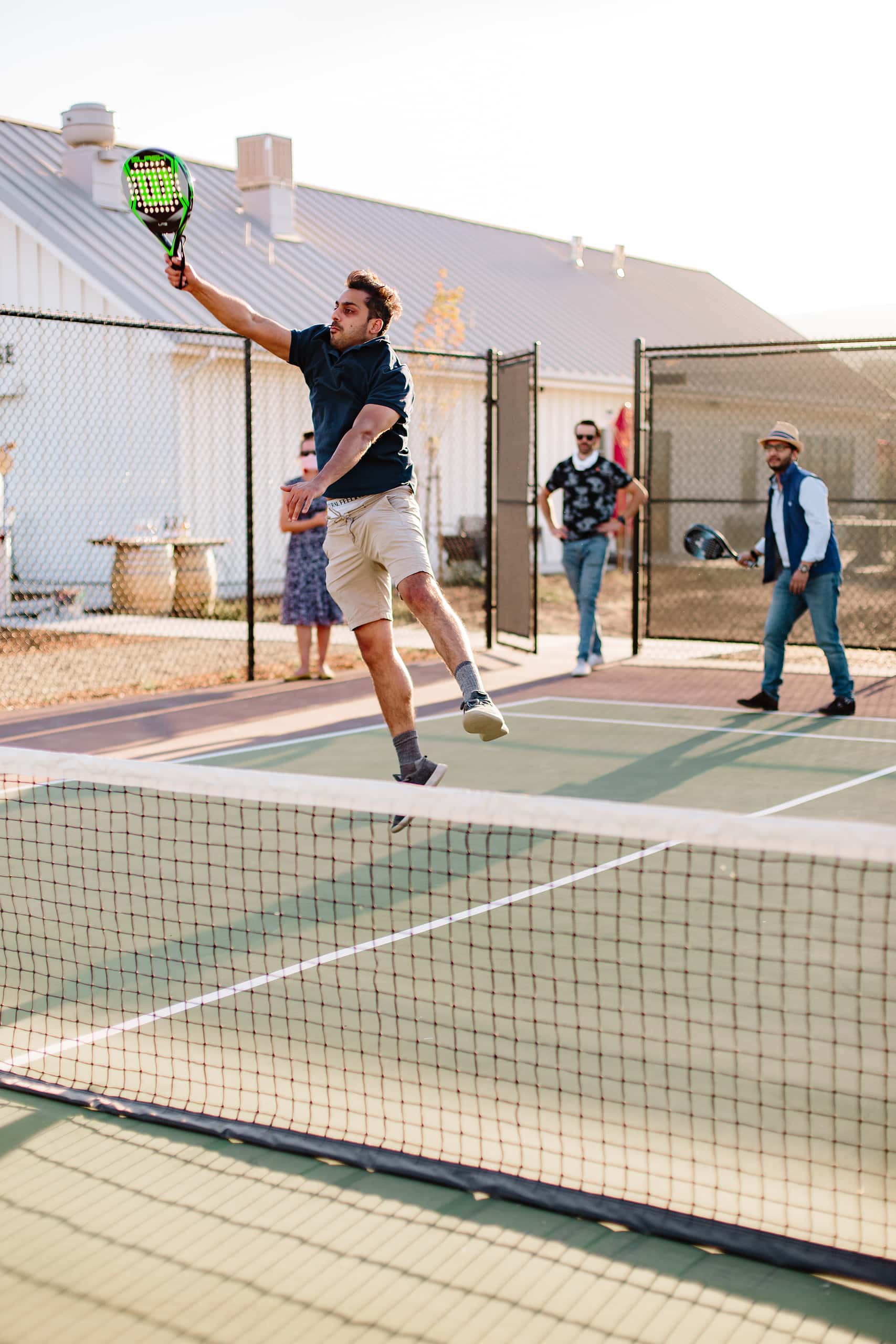 A group of friends on the pickleball court, a man jumping to hit the ball in the foreground, his friends blurred in the background. 