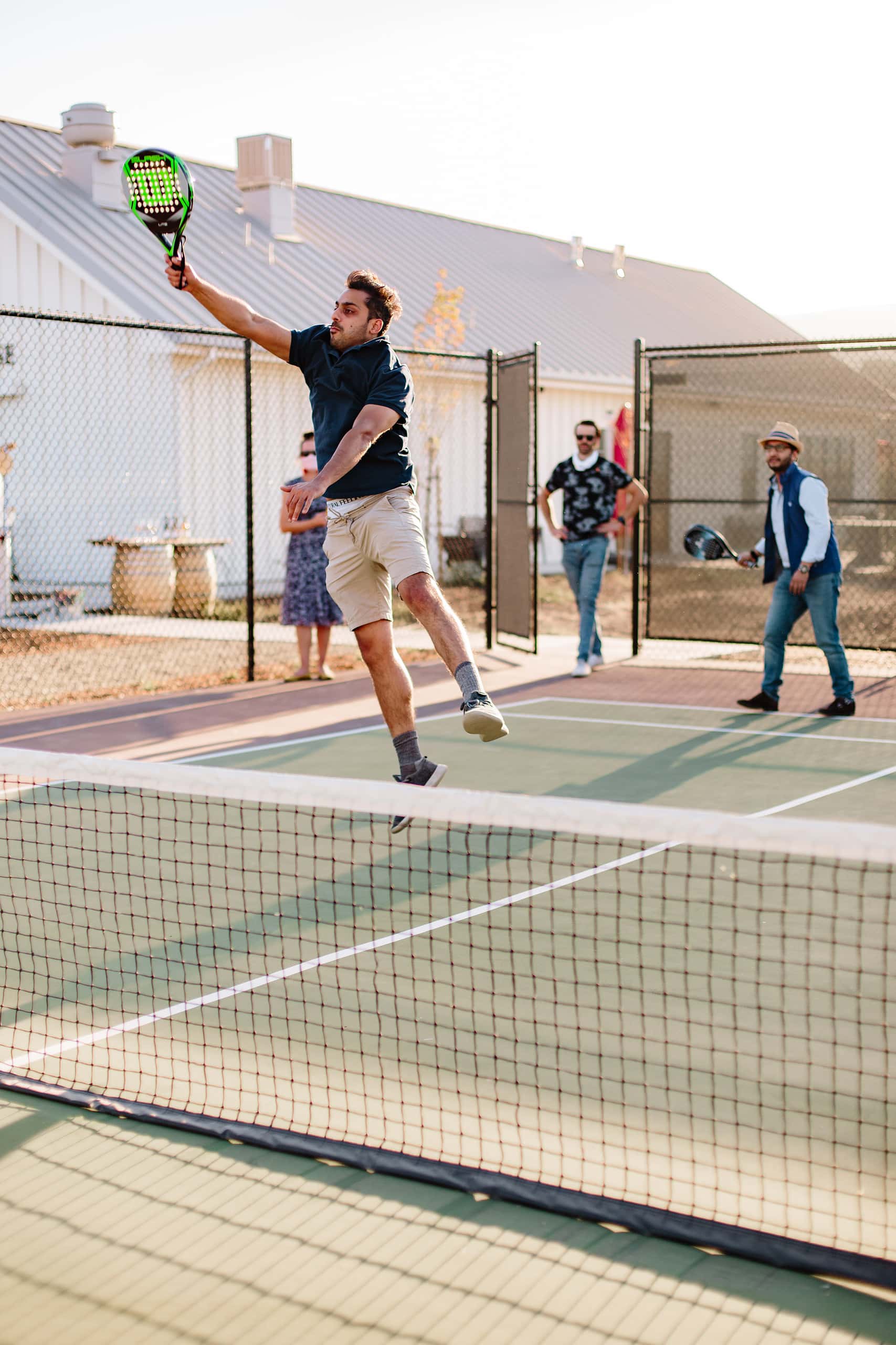 A group of friends on the pickleball court, a man jumping to hit the ball in the foreground, his friends blurred in the background.