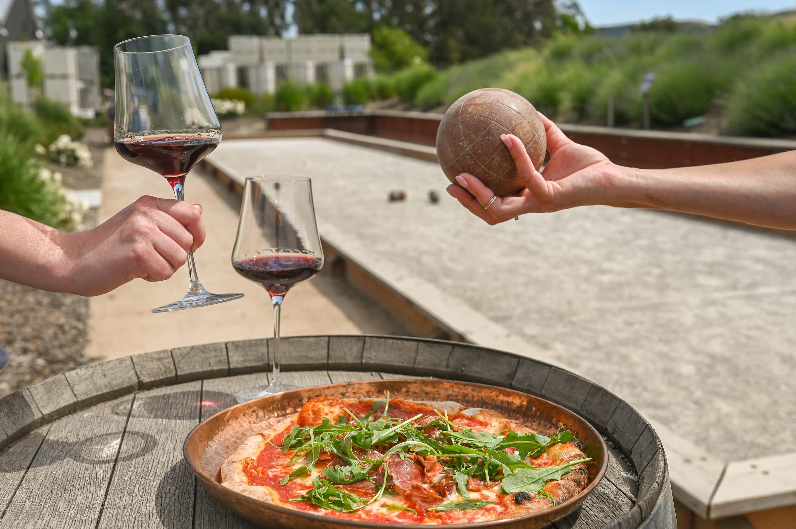 Woman holding bocce ball, man holding glass of red wine, set over an over fired pizza in front of Bocce area.