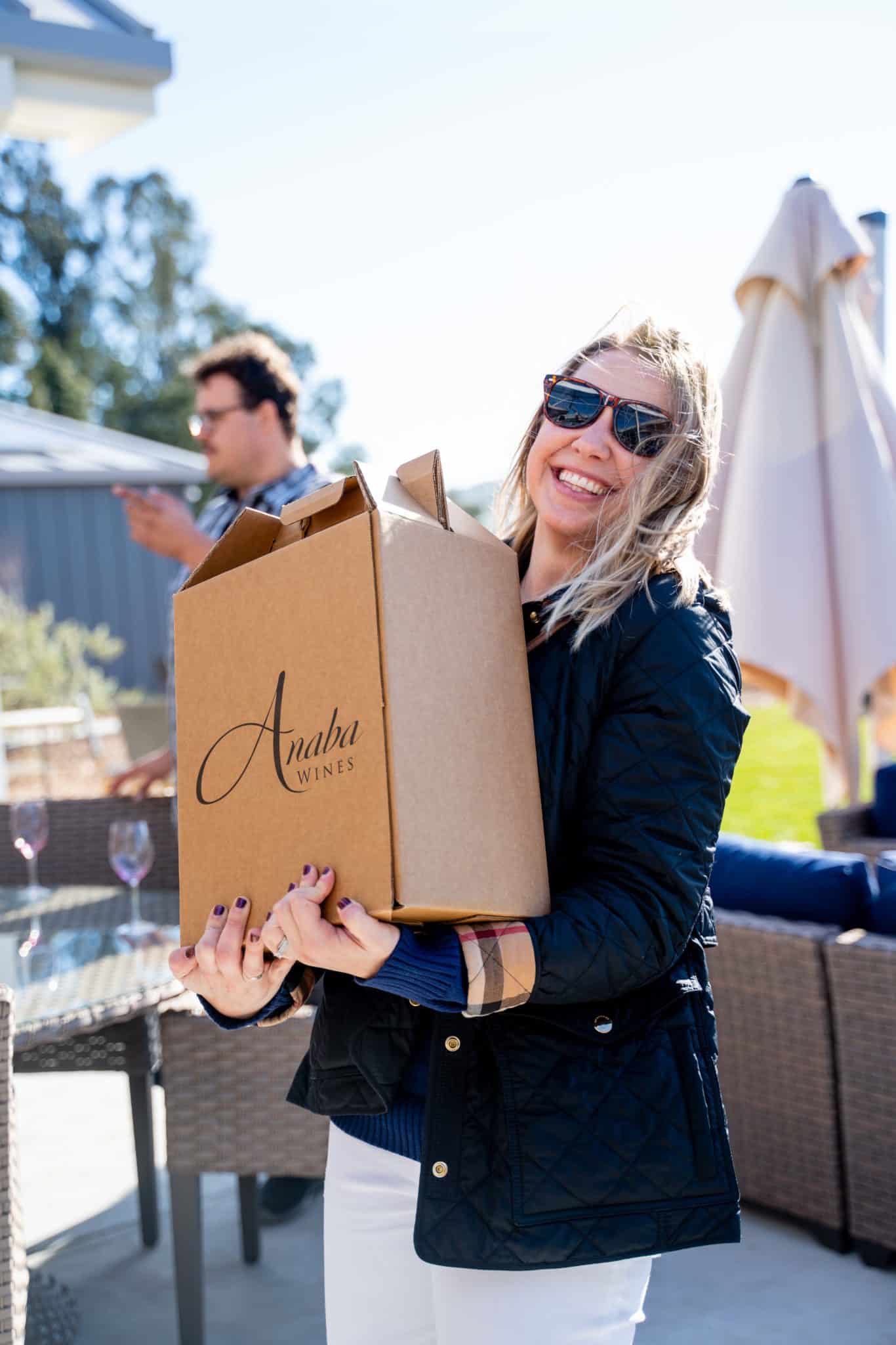 A woman in the foreground, smiling whilst carrying a Anaba Wines box. 