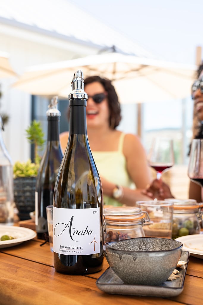 A group of friends sat drinking Anaba wine together, blurred in the background, a bottle of Anaba Turbine White wine placed in the foreground. 