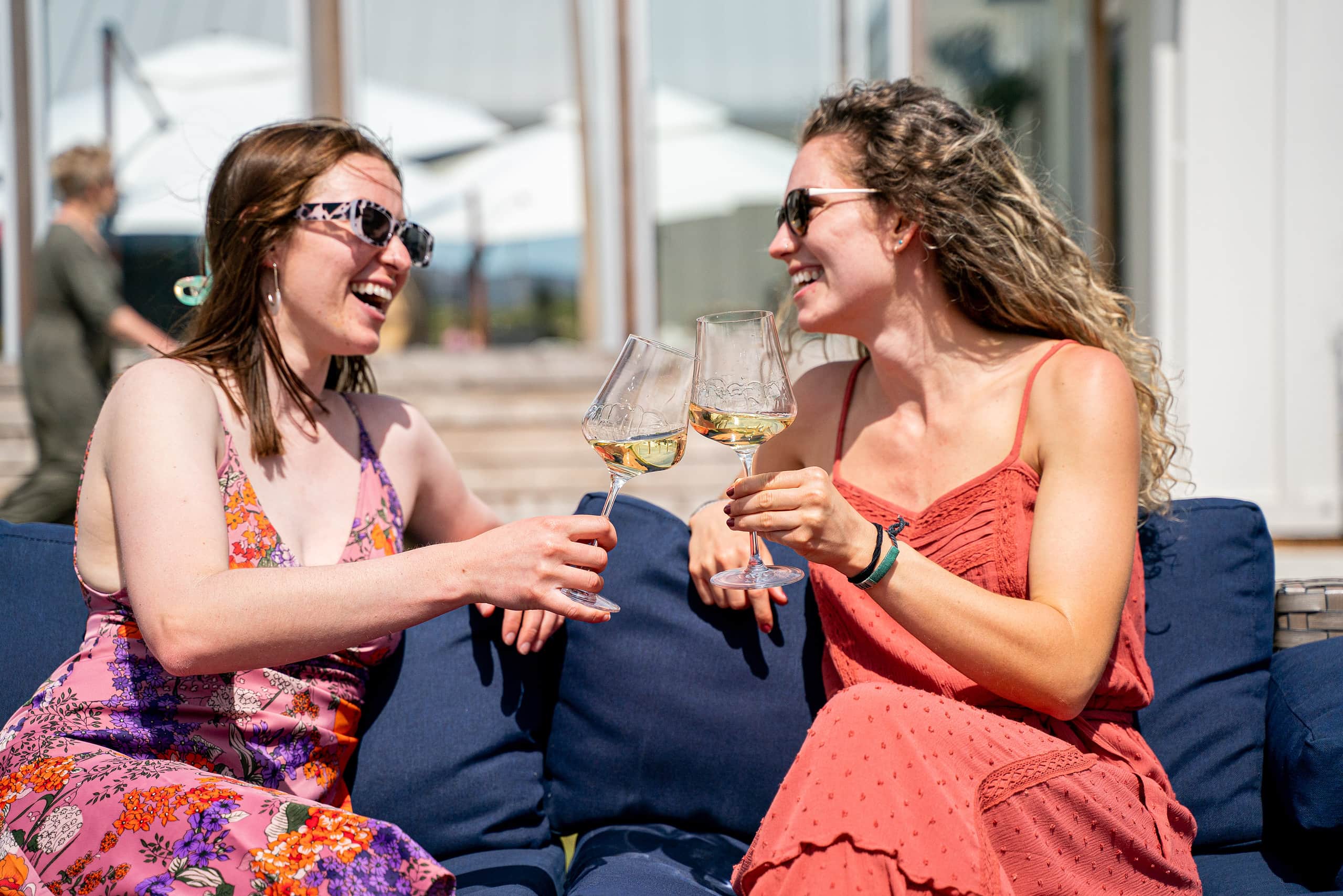 Two girls sat on a blue couch drinking wine together and laughing. 