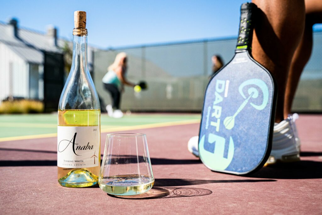 Bottle of Anaba Wines and stemless glass on ground of pickleball court next to person sitting with their racket.