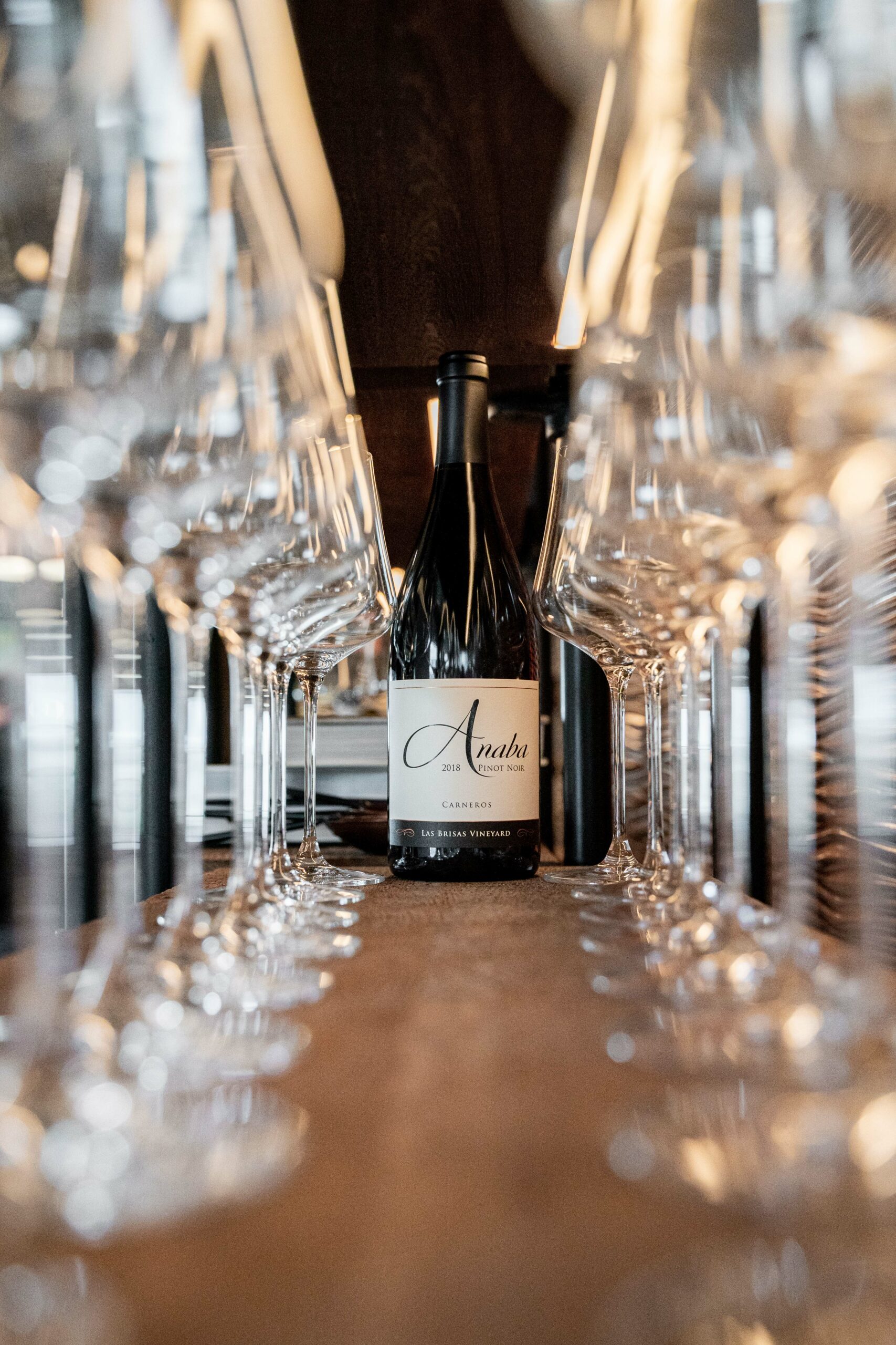 A low angle shot of Anaba Pinot Noir centred in the mid-ground, with rows of blurred wine glasses in the foreground, lining either side of the image, leading up to the bottle of wine. 