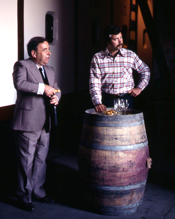 A young John Sweazey on stage with another man, leaning against a wine barrel while the man opens a bottle of wine. Both men are well lit in the foreground, while the background is blurred and dark. 
