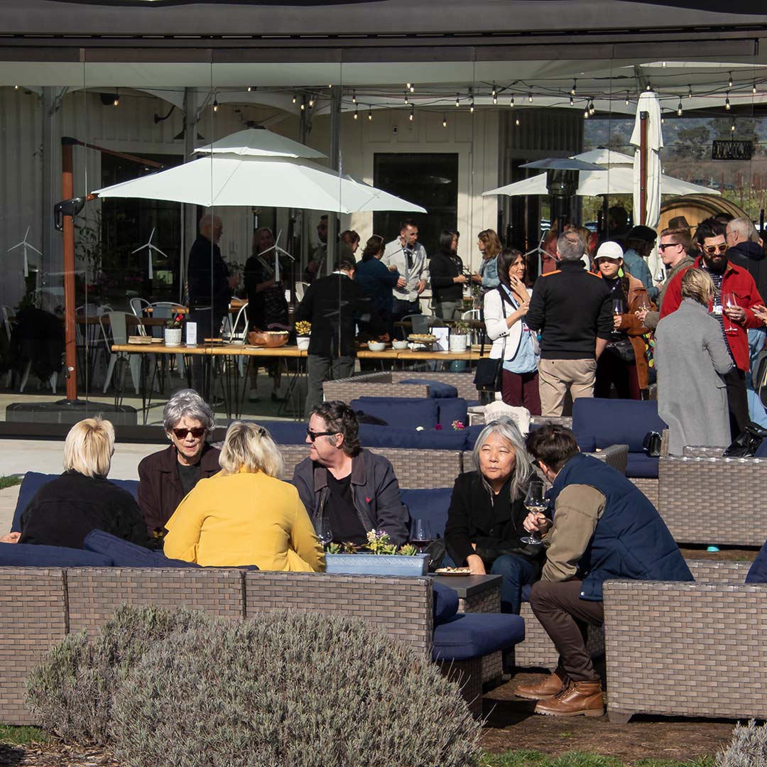 A wide angle shot of people relaxing, drinking wine, whilst sitting or standing around tables. 