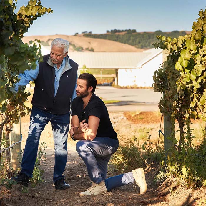 John Sweazey and John Michael standing between two vines in the J McK Estate Vineyard, blooming vines in the foreground, a building in the mid-ground, with hills of trees in the background. 