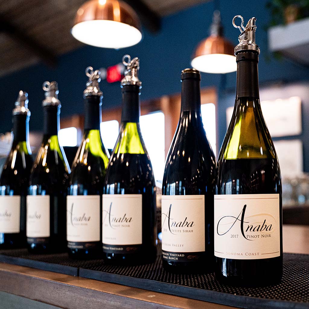 A line of six Anaba wine bottles placed on a table, inside, with a blurred kitchen in the background. 