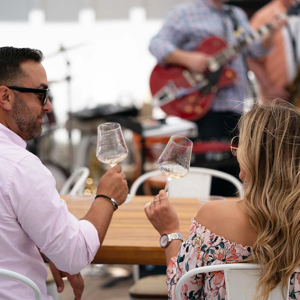 A man and a woman sat in the foreground, drinking white wine, with a band performing in the blurred background. 