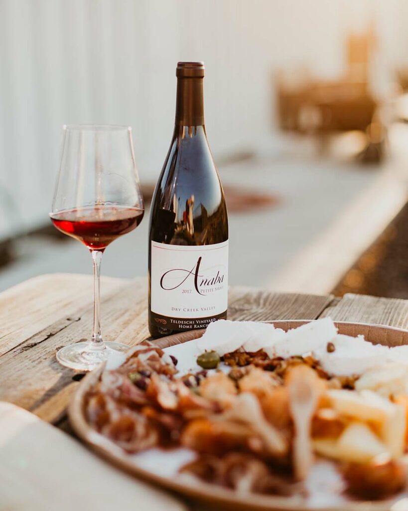 A platter of food blurred in the foreground, a bottle and glass full of Petite Sirah in the mid-ground. 