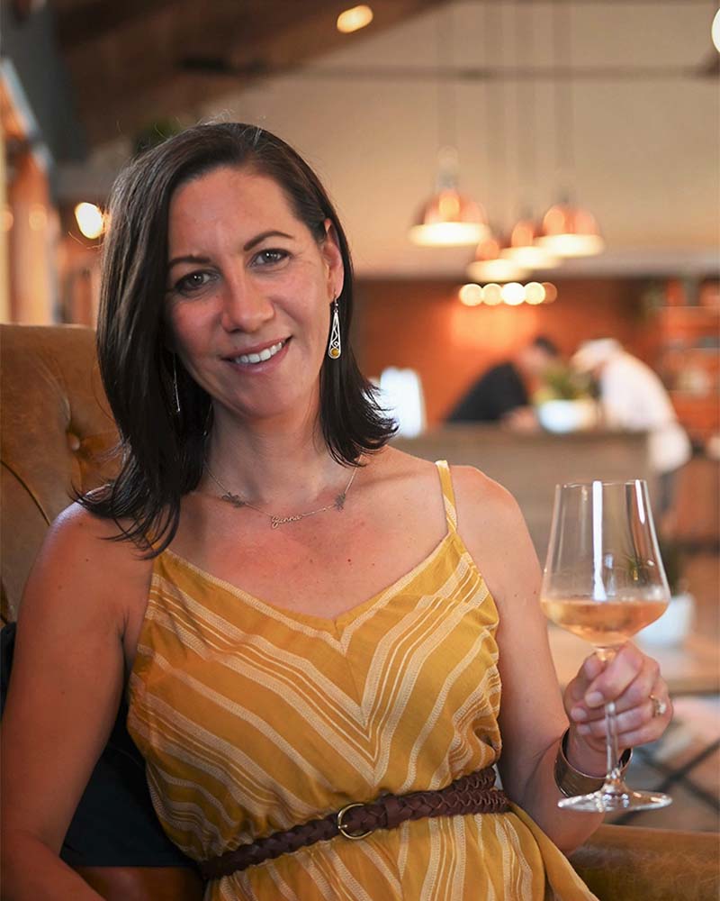 Alaina Murphy smiling, holding a glass of rosé in the foreground, a blurred tasting room in the background.