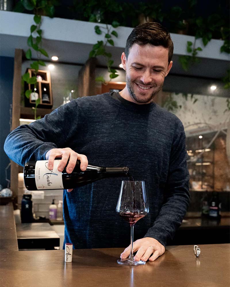 Ben Curley centred in the foreground, pouring Anaba red wine with his right hand, his left hand steadying the glass, with the Anaba tasting room in the background. 