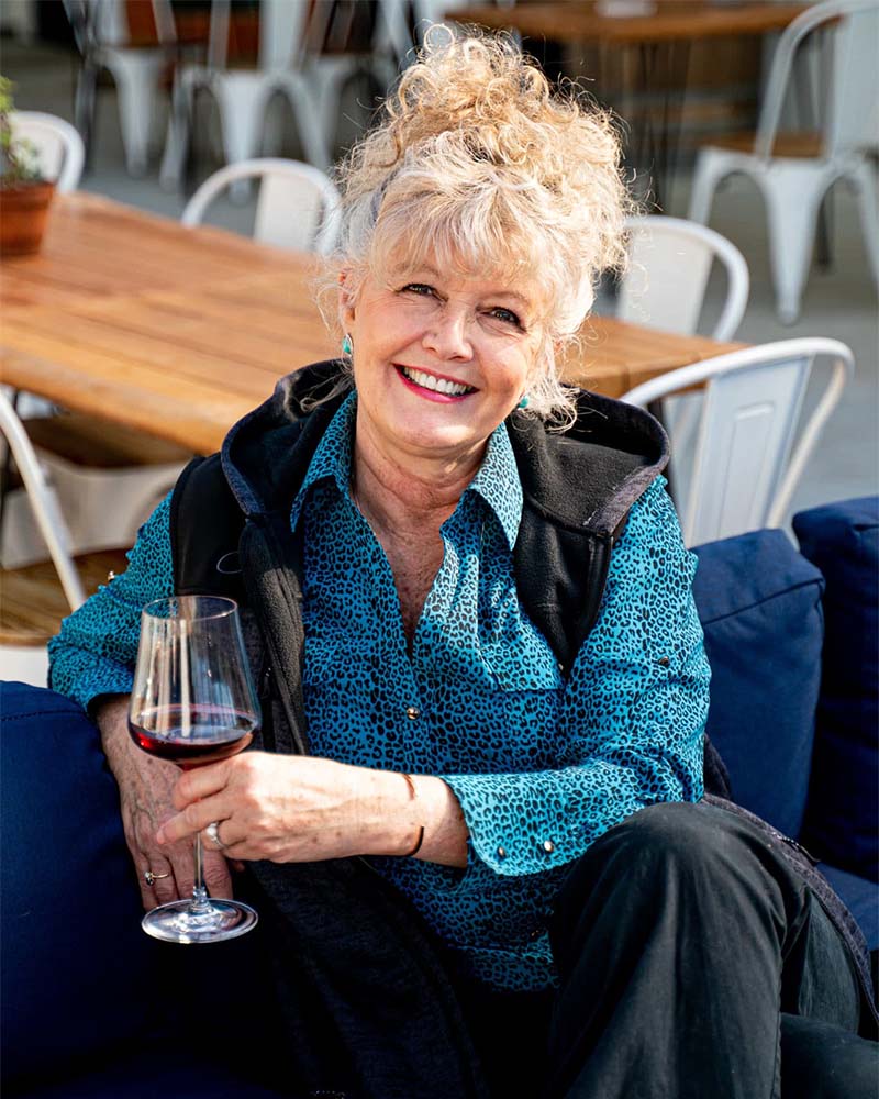 Casandra Knox seen in the foreground, smiling, holding a glass of red wine whilst sat on a blue couch, sets of tables and chairs arranged in the background. 