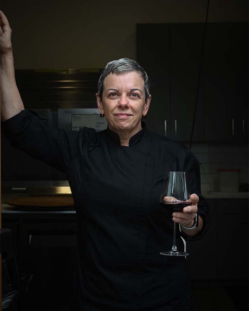 Chef Maria centred in foreground, her right arm raised, her left holding a glass of red wine, a dim kitchen seen in the background. 