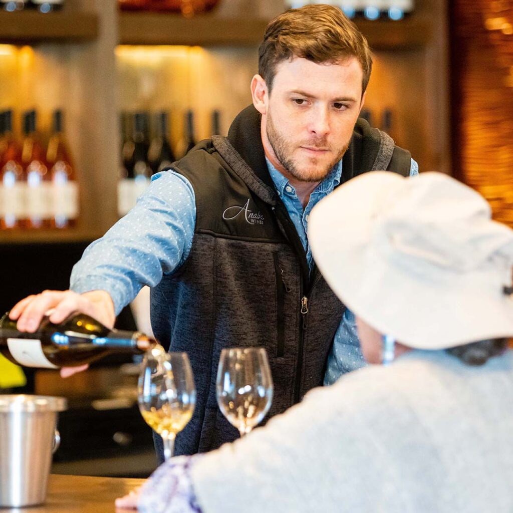 An employee of the Anaba vineyard pouring glasses of wine to a customer in the blurred foreground, the background showing a cabinet with rows of Anaba wine. 