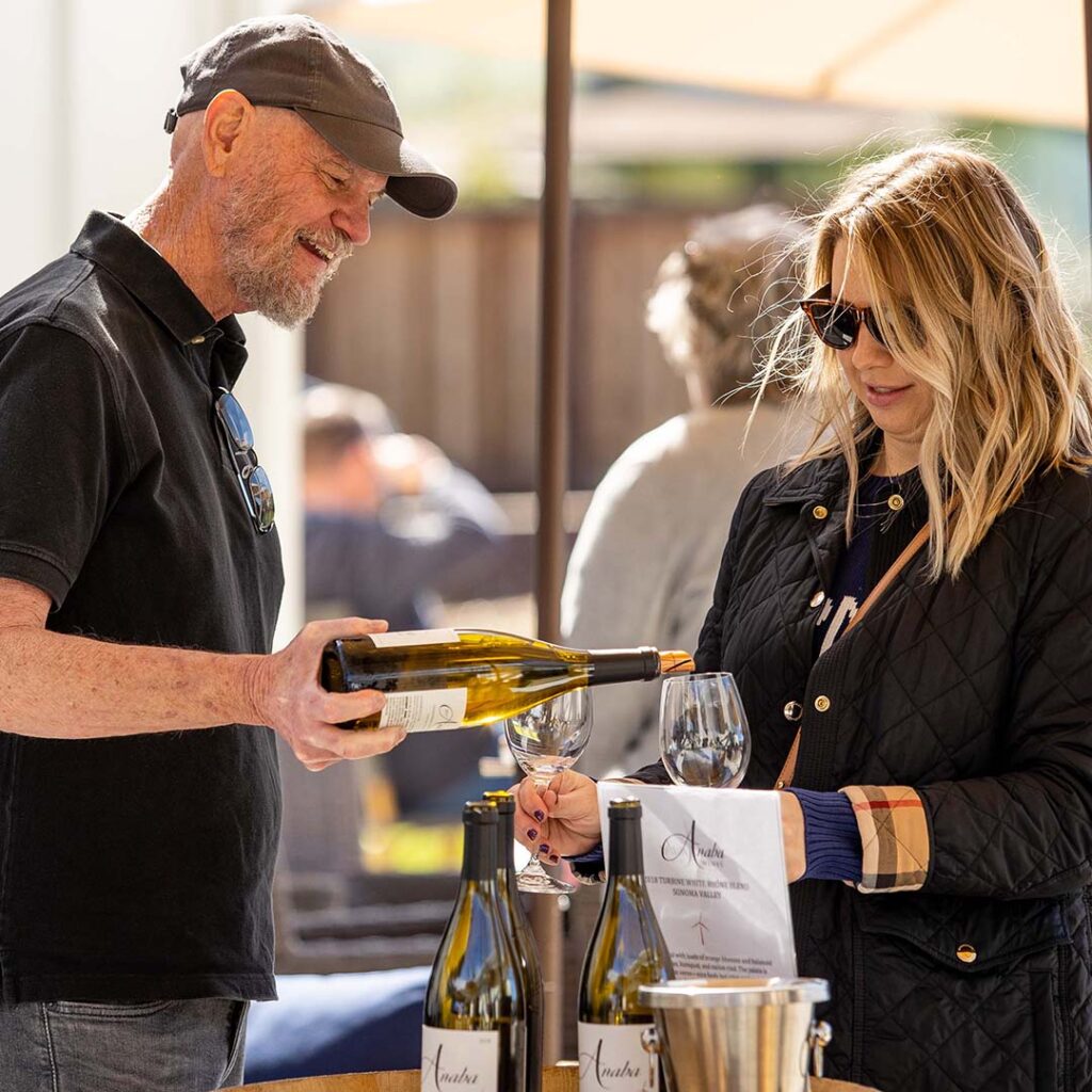 A man and a women stood in the foreground, pouring two glasses of Anaba white wine, other customers relaxing, blurred in the background. 