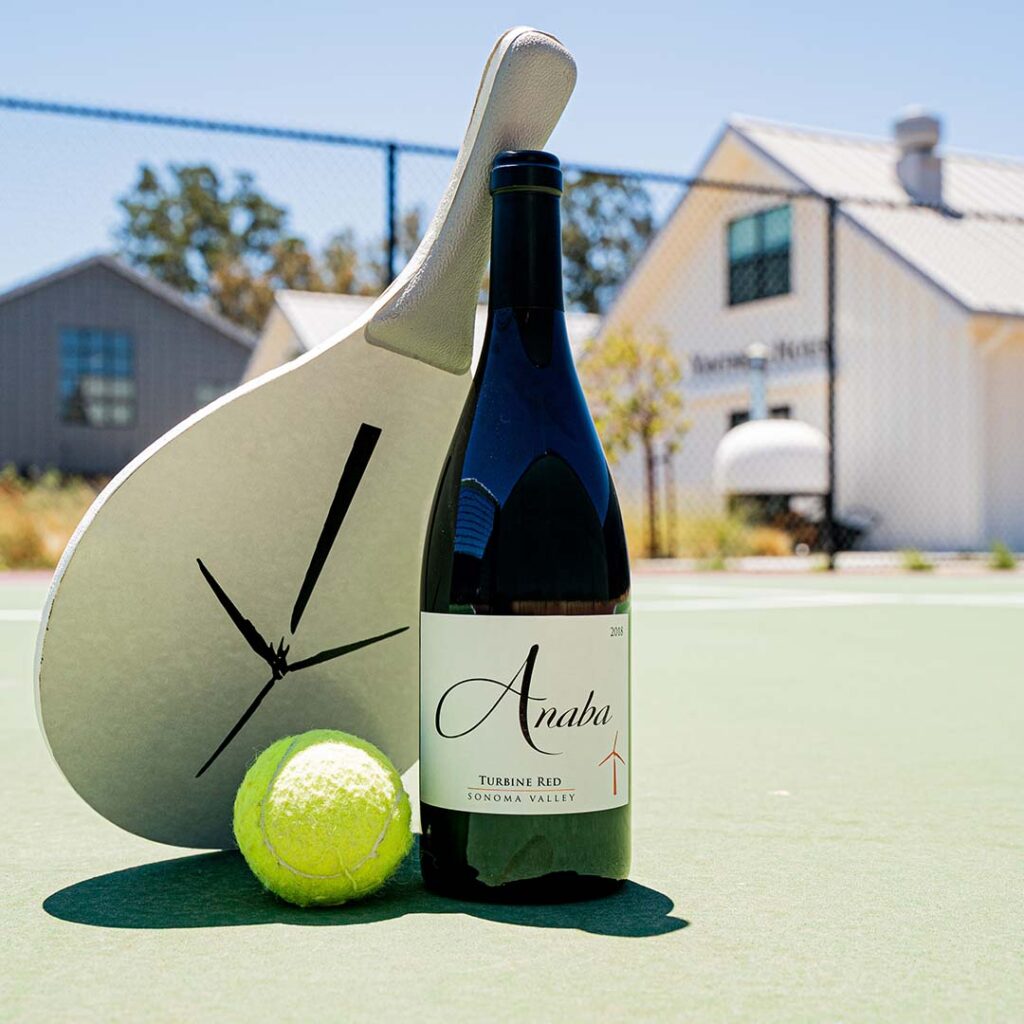 A bottle of Anaba Turbine Red wine placed on a pickleball court, with a bat and ball positioned against the wine bottle, the buildings of the vineyard seen blurred in the background. 