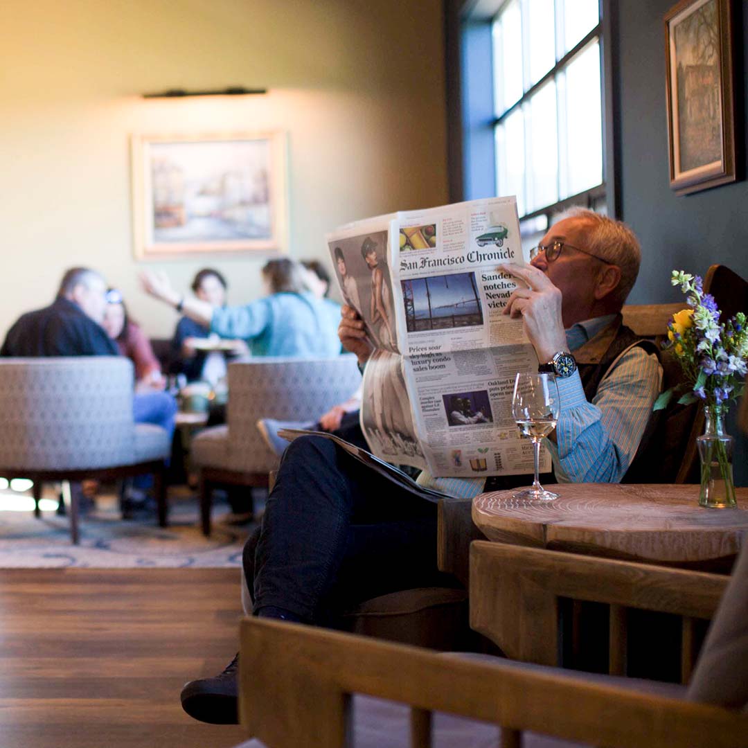 A man sat on an armchair reading the San Francisco Chronicle, with a glass of wine on the table beside him in the foreground, the blurred background showing a large group of friends sat, talking, in armchairs. 