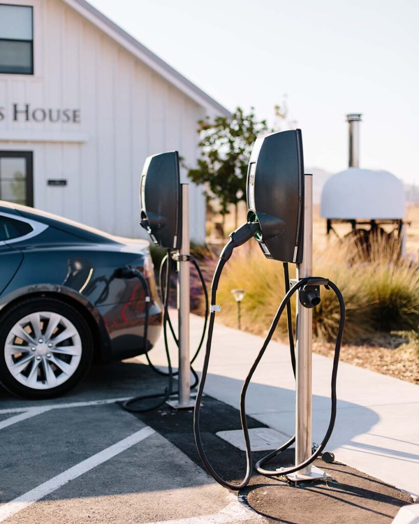 Two electric car charges centred in the foreground, a black car being charged from one of them, a blurred building and desert shrubbery in the background.