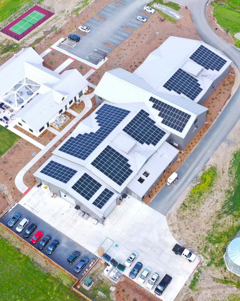 A birds eye view of the Anaba Wines winery, showing a large grey building with solar panels lining the each section of the roof, small parking lots on either side of the building, and a smaller white building connecting the three sections. 