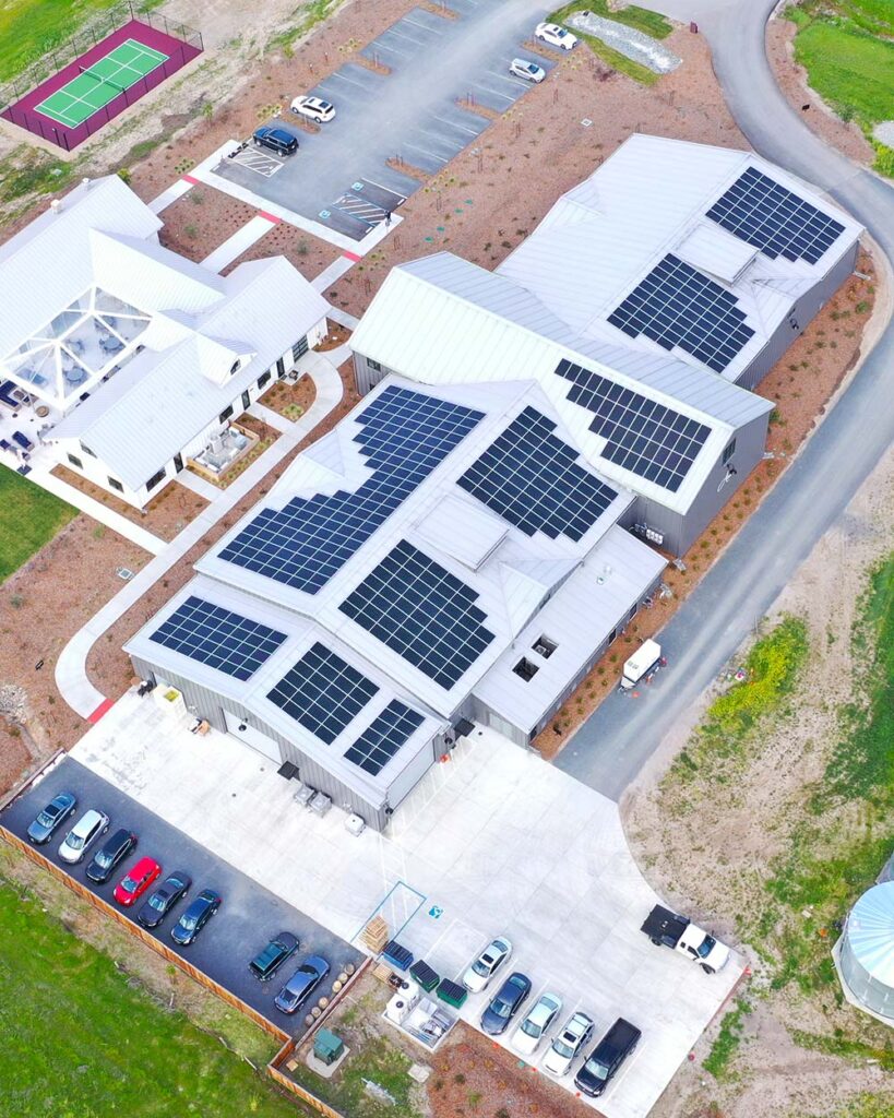 A birds eye view of the Anaba Wines winery, showing a large grey building with solar panels lining the each section of the roof, small parking lots on either side of the building, and a smaller white building connecting the three sections.