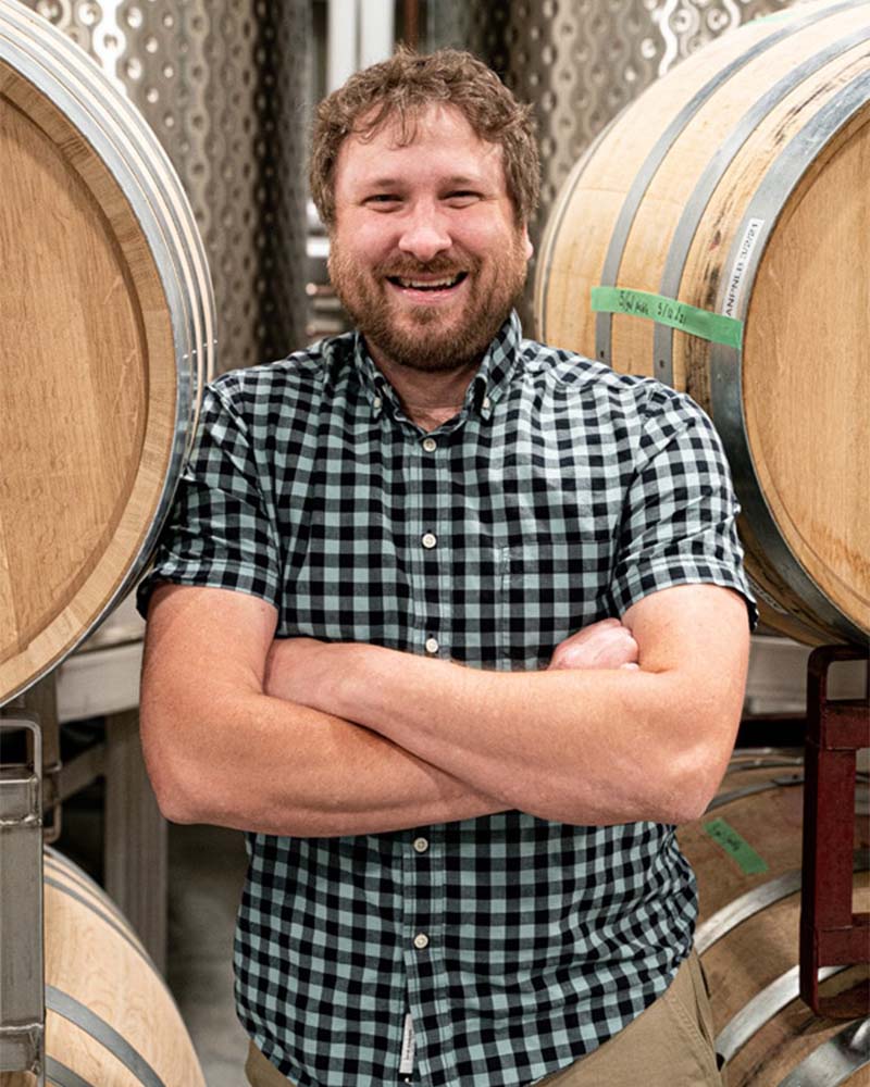 Ian McClellan centred between four wine barrels, smiling with his arms crossed.
