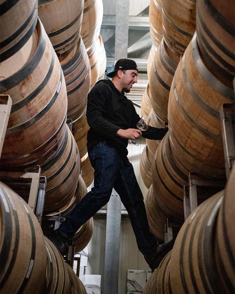Jesse Perez smiling, holding a wine glass, whilst balanced between two rows of wine barrels, each foot resting on a barrel on either side of him. 