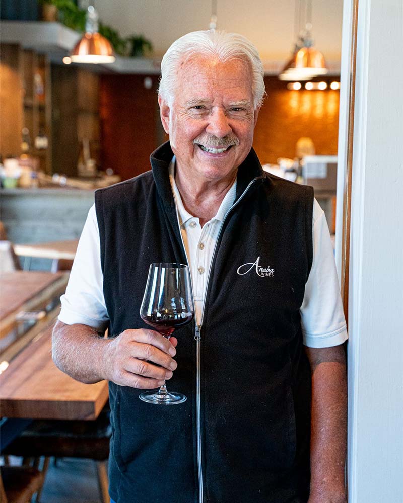 John Sweazey smiling, holding a glass of red wine in the foreground, a blurred tasting room in the background. 