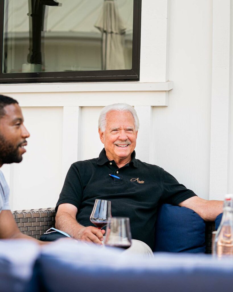 A smiling John Sweazey sitting on a blue couch, drinking a glass of red wine in the midground, the blurred foreground showing another man sat with Sweazey. 