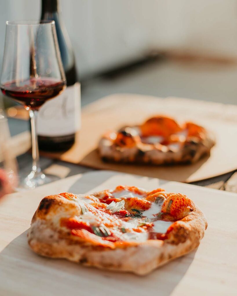 A pizza placed on a wooden platter in the foreground, another pizza and wine blurred in the background. 