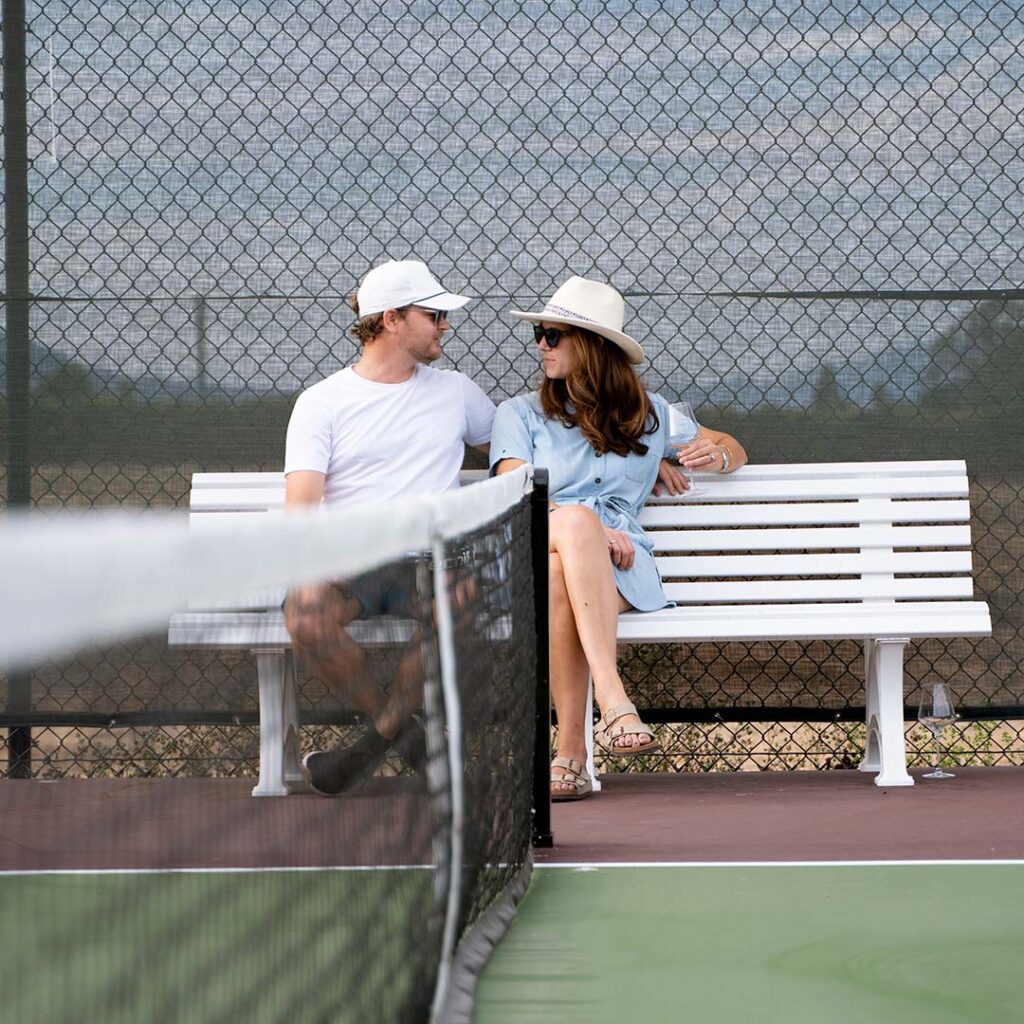 In the mid-ground a man and a woman are sat together on a bench in the pickleball court, the net blurred in the foreground. 