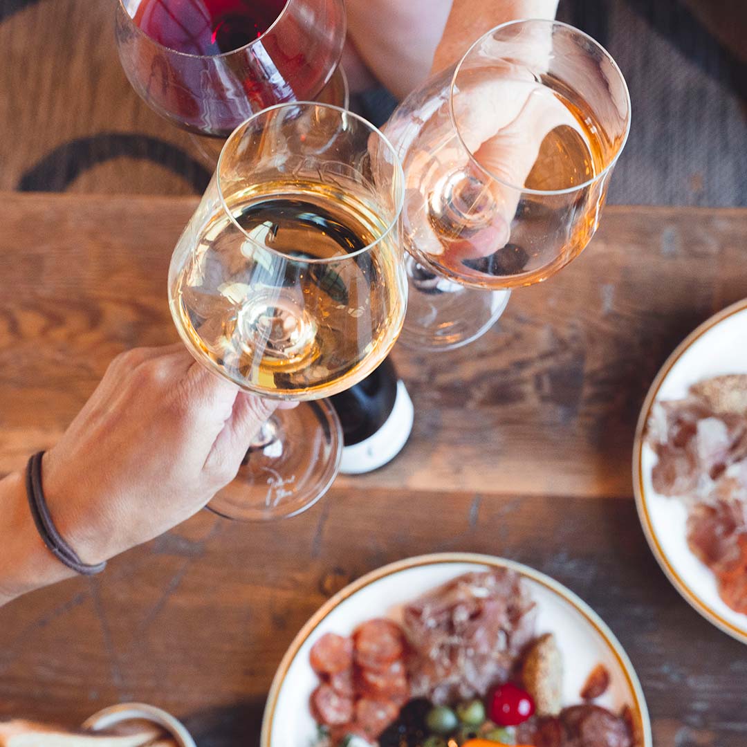 A birds eye view of three wine glasses clinking glasses over a dark wooden table, with platters of food in the background.