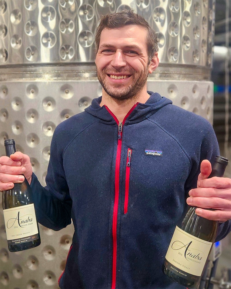 Winery Production Manager Brian Mascia wearing a blue hoodie, holding two bottles of Anaba Wine and smiling at camera.