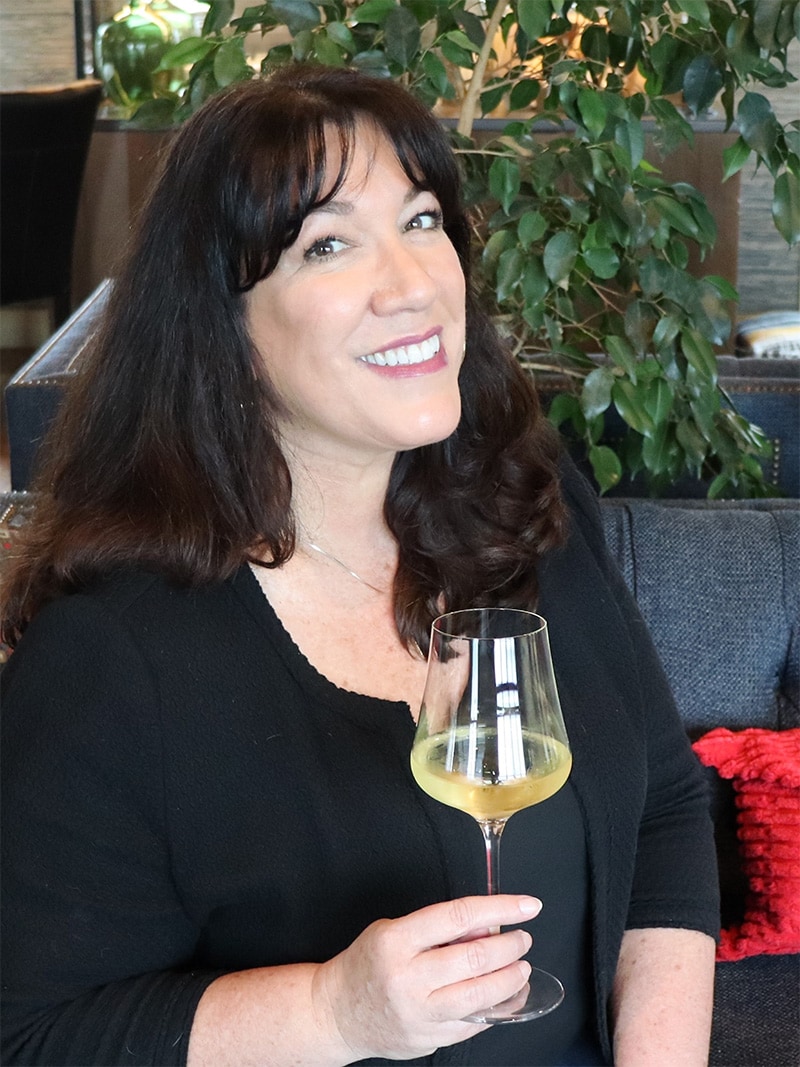 Michelle Hogan sitting on a couch in the Anaba Tasting Room, holding a glass of white wine while she smiles at camera.