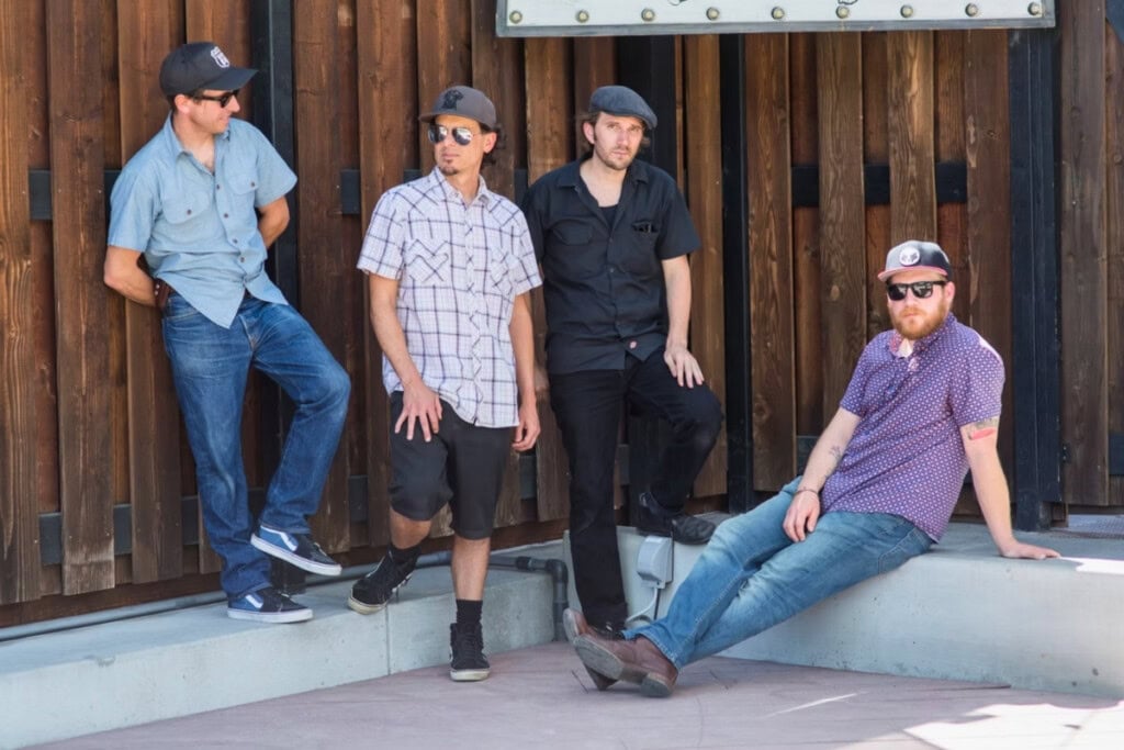 Four young men pose outdoors against a wooden wall. They wear casual clothing and hats. One leans against the wall, two stand, and one sits on a ledge.