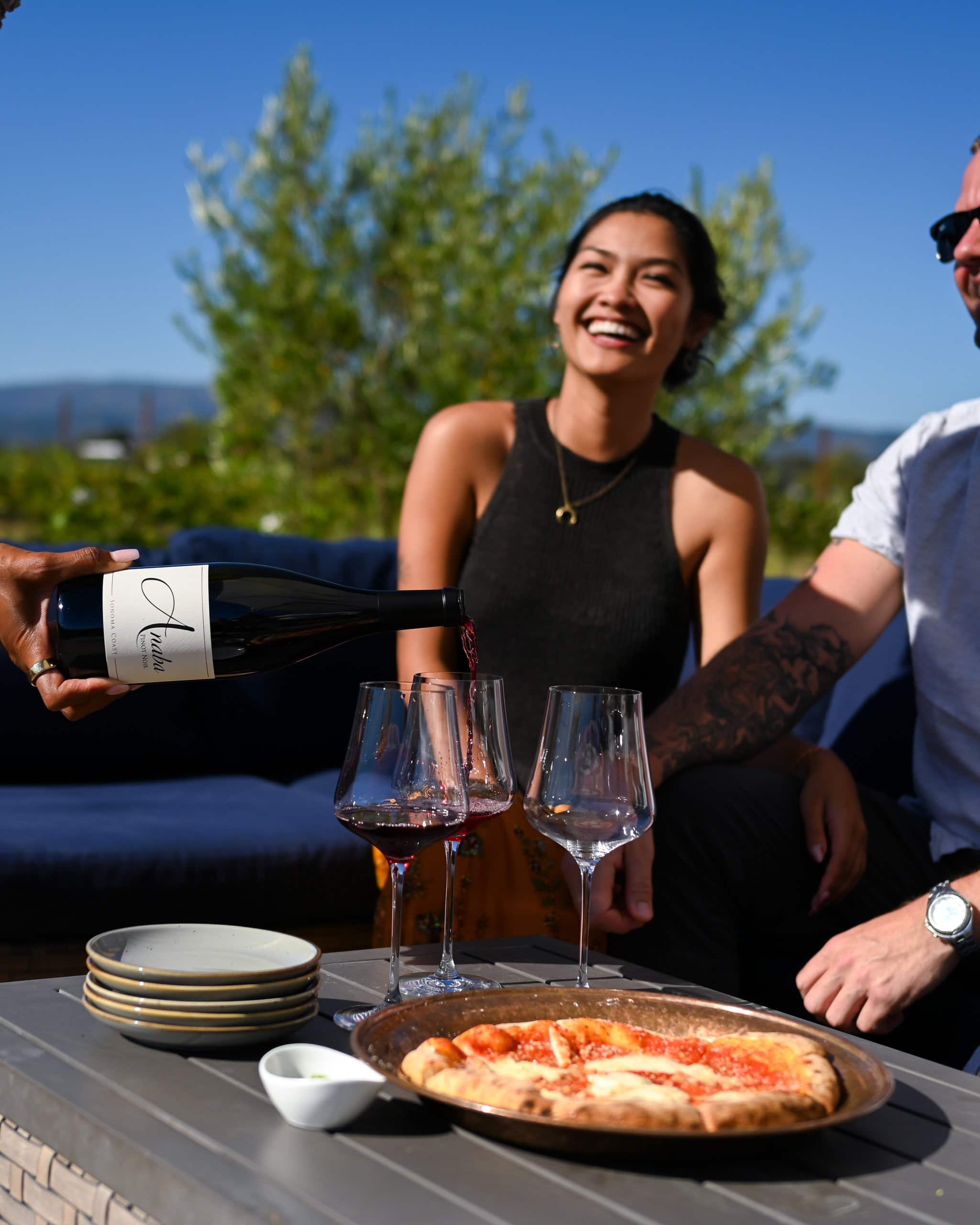 Two friends sitting outside at Anaba Tasting Room. They are enjoying wine and pizza. Someone off camera is pouring red wine into glasses