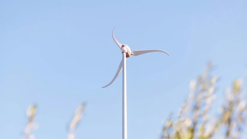 A Skystream wind turbine stands against a clear, light blue sky, with blurred foliage visible at the bottom of the frame.