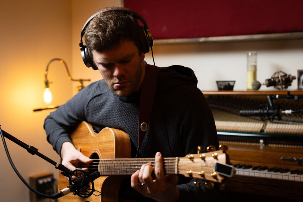 Picture of man wearing headset playing a guitar with a keyboard behind
