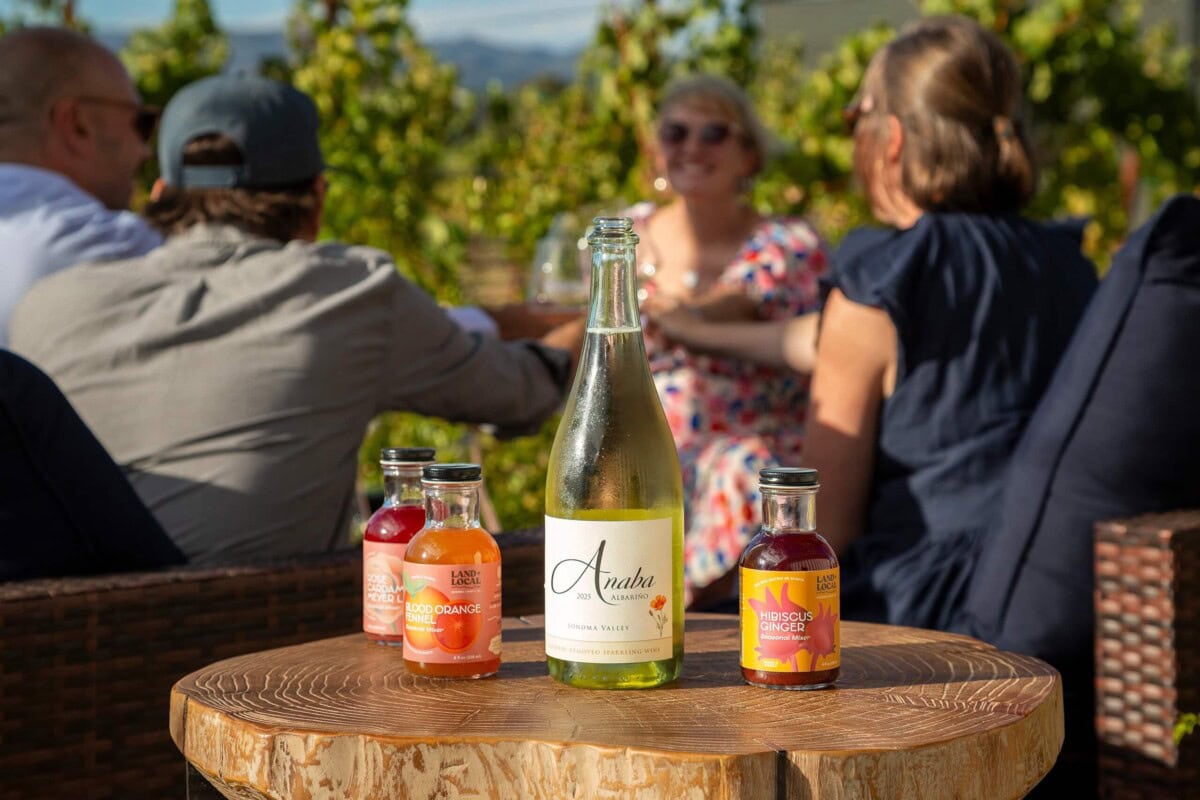 Anaba Wines bottle on a rustic wooden table with attendees in vineyard lounge setting in the background.